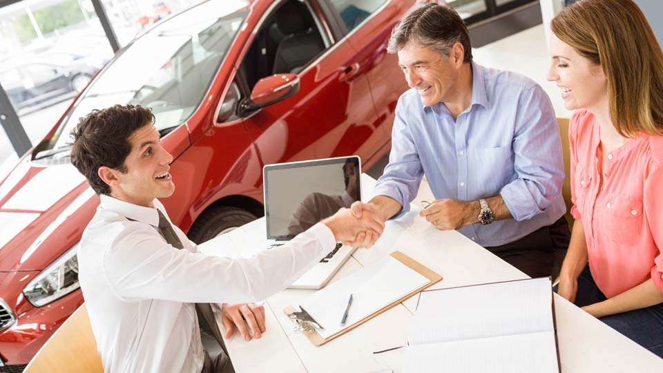 Ein Autoverkäufer schüttelt den Kunden die Hand in einem Autohaus in Bad Hersfeld, im Hintergrund ist ein rotes Auto zu sehen und auf dem Tisch liegen Unterlagen.